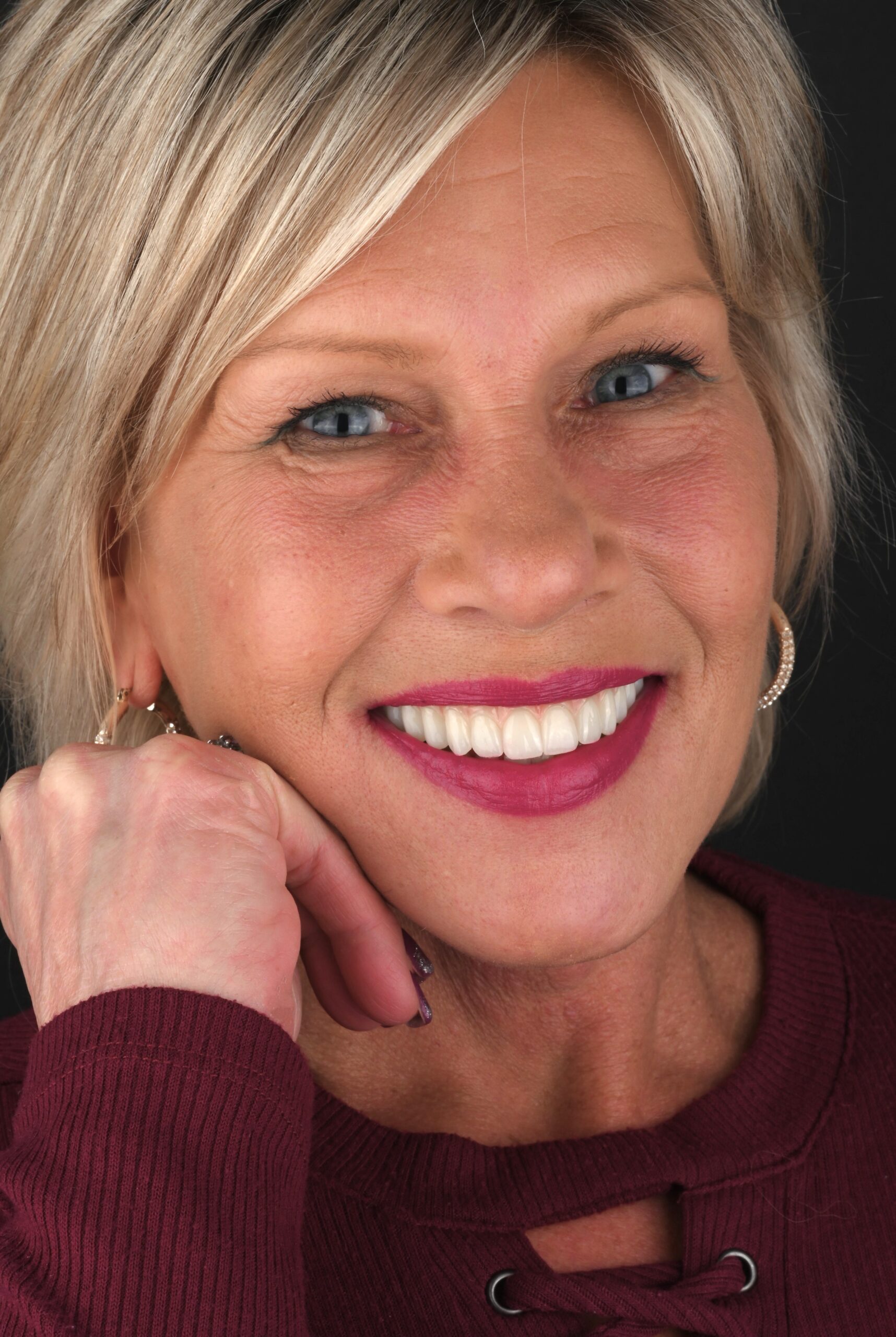 Close-up of a smiling woman with blonde hair and blue eyes, wearing a burgundy sweater and resting her chin on her hand.