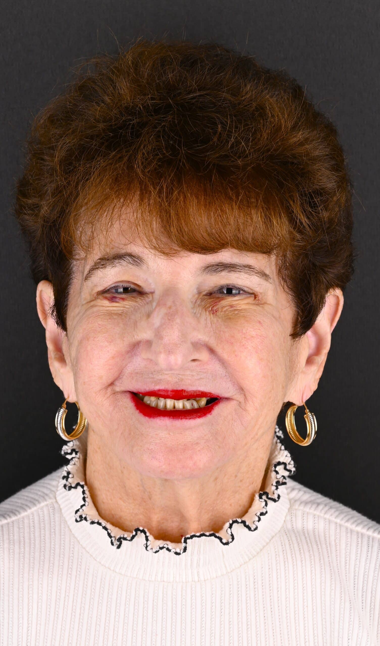 Smiling older woman with short brown hair, gold hoop earrings, and a white ribbed sweater against a dark background.