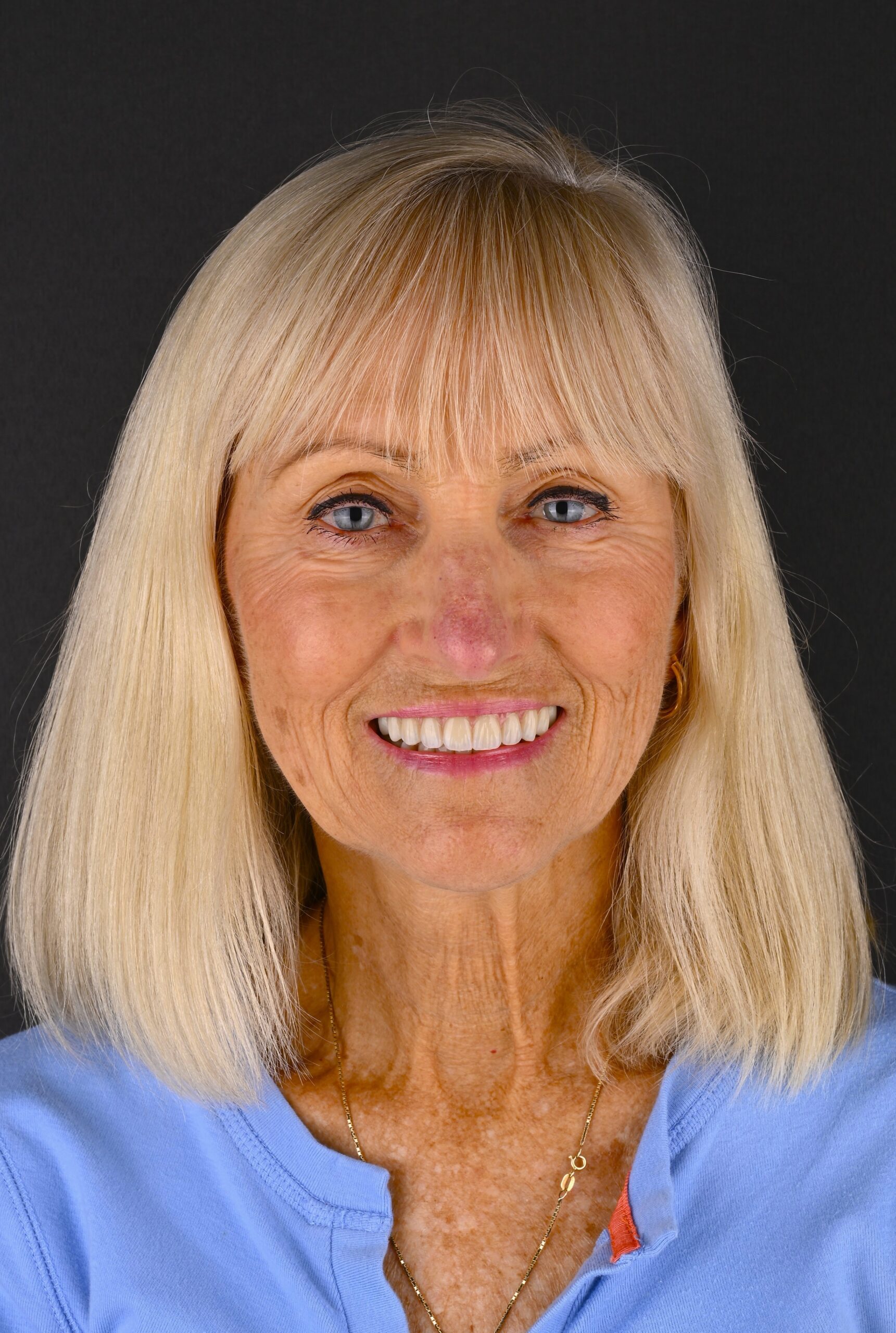 Smiling close-up of an older woman with long blonde hair and blue eyes, wearing a light blue top against a dark background.