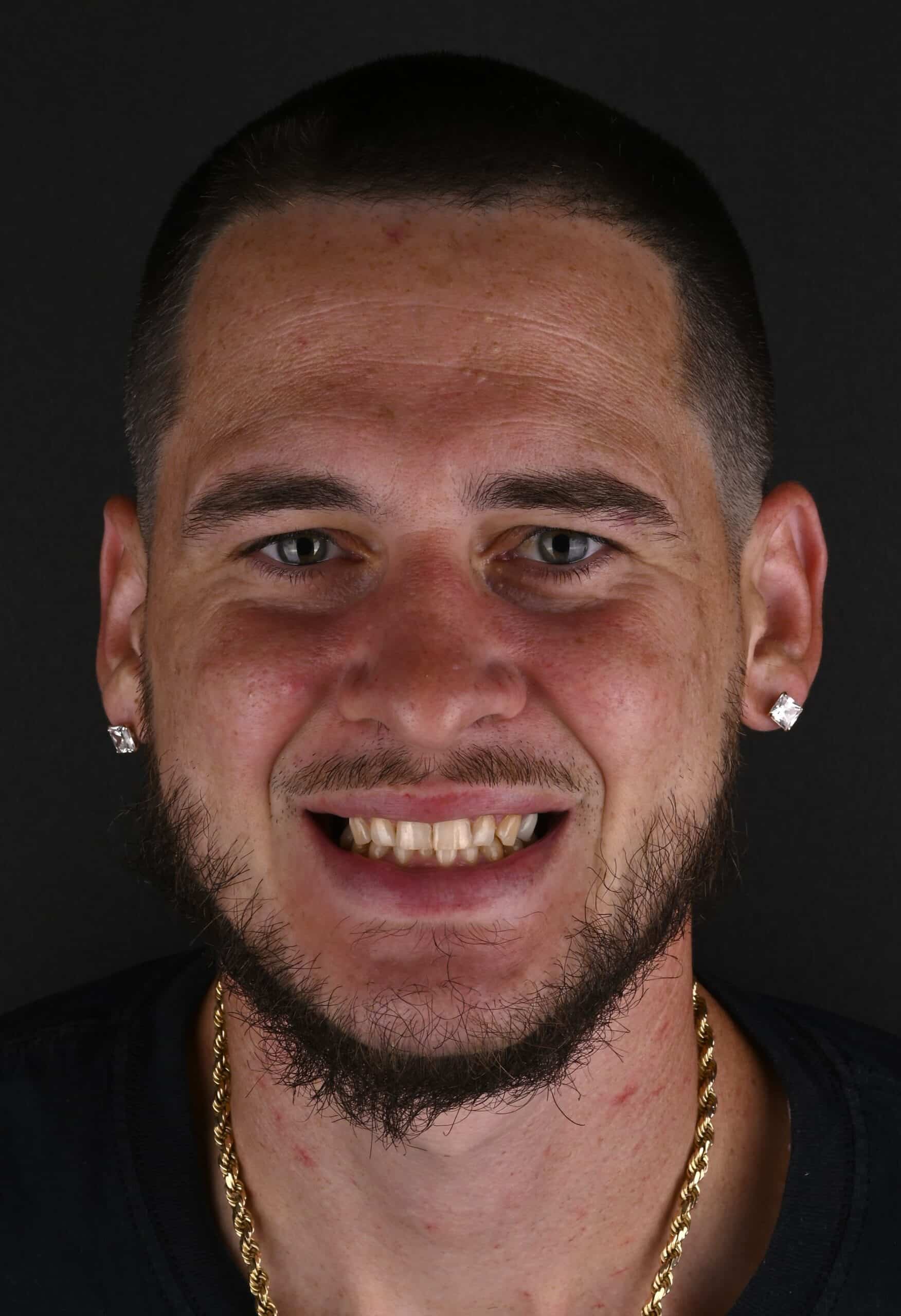 Close-up of a smiling man with short dark hair and a beard, wearing diamond earrings and a gold chain against a black background.