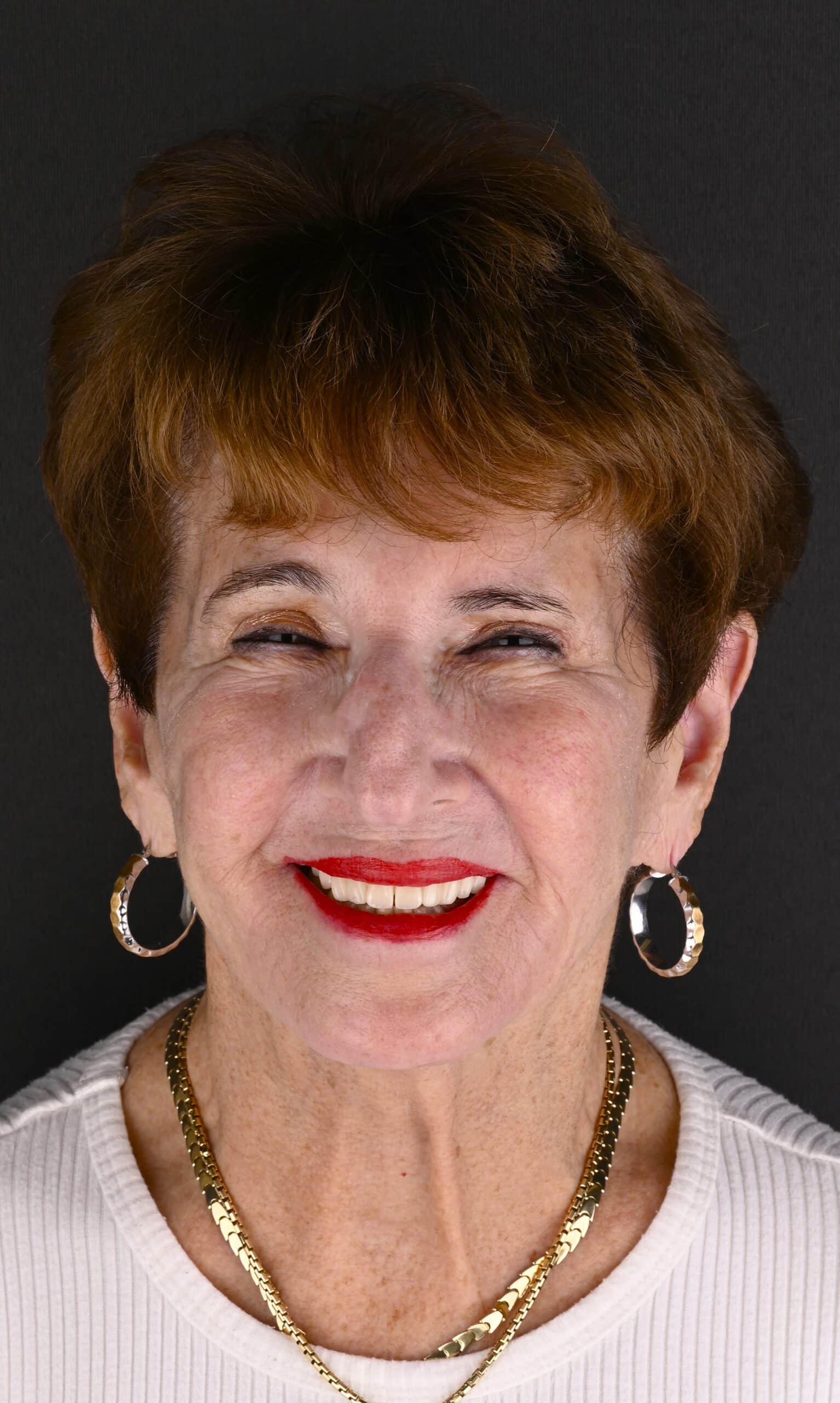 Smiling older woman with short brown hair, red lipstick, hoop earrings, and a gold chain against a dark background.
