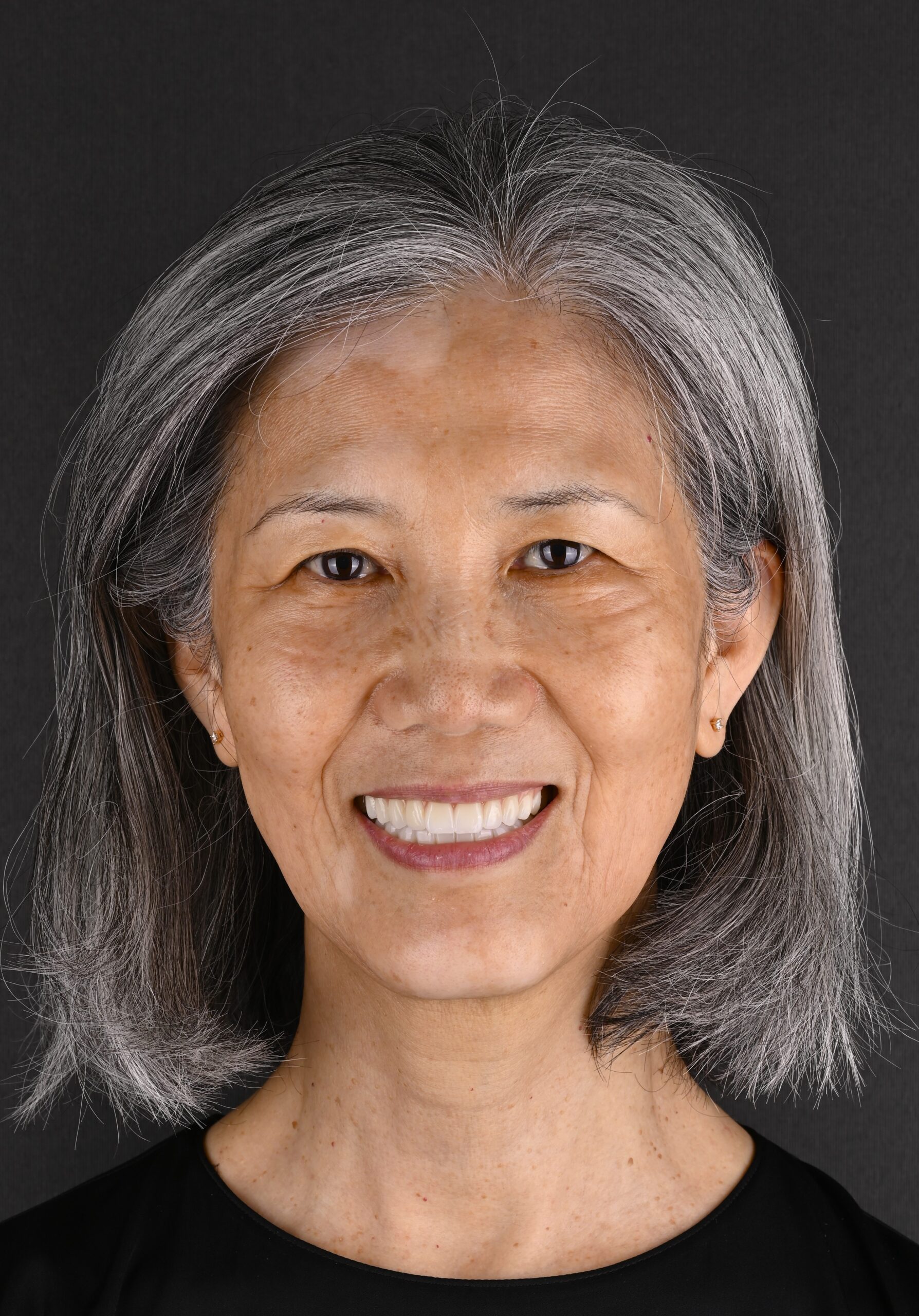Close-up portrait of a smiling woman with silvery gray hair against a dark background.