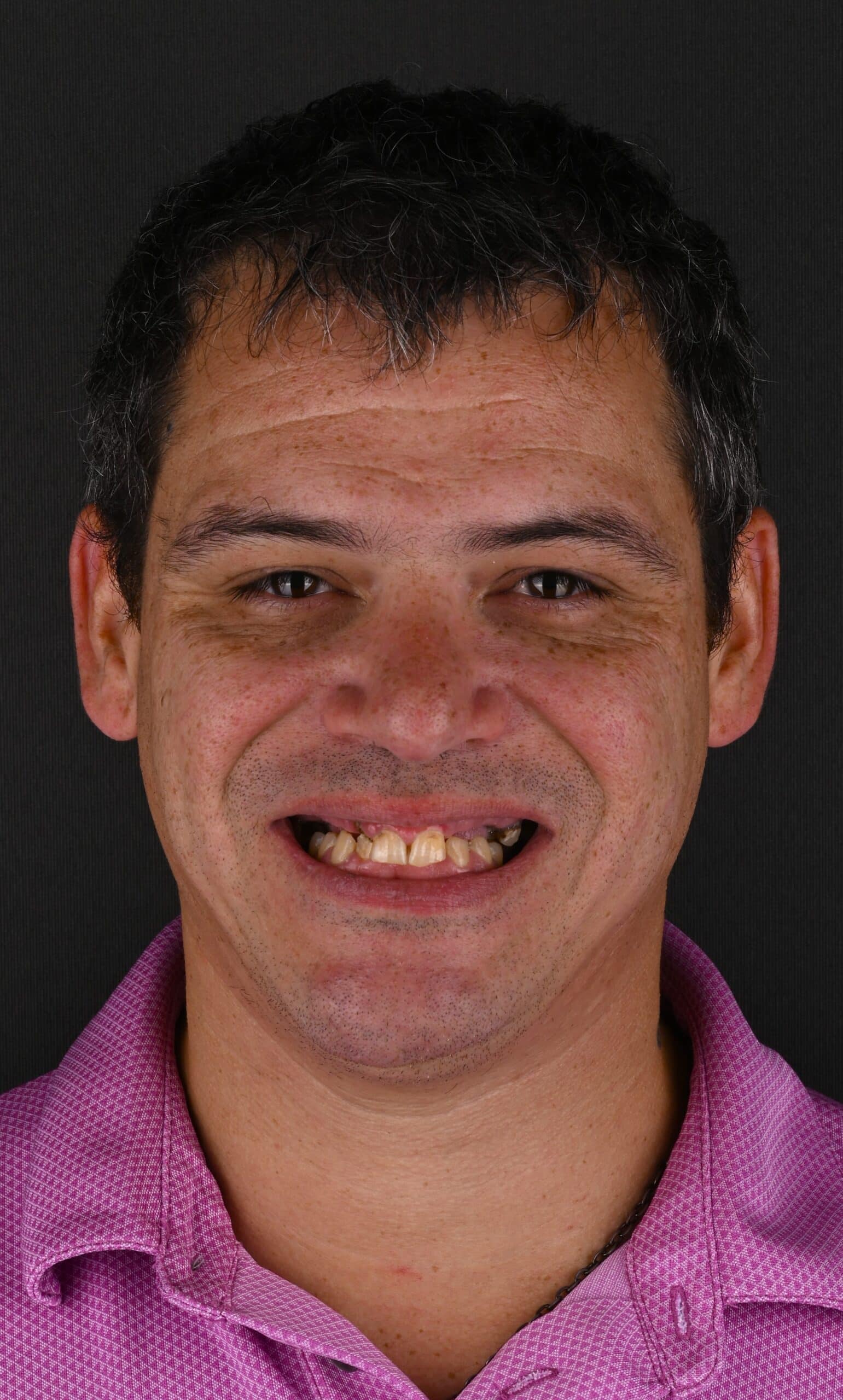 Close-up portrait of a smiling man with short dark hair, wearing a pink shirt against a dark background.