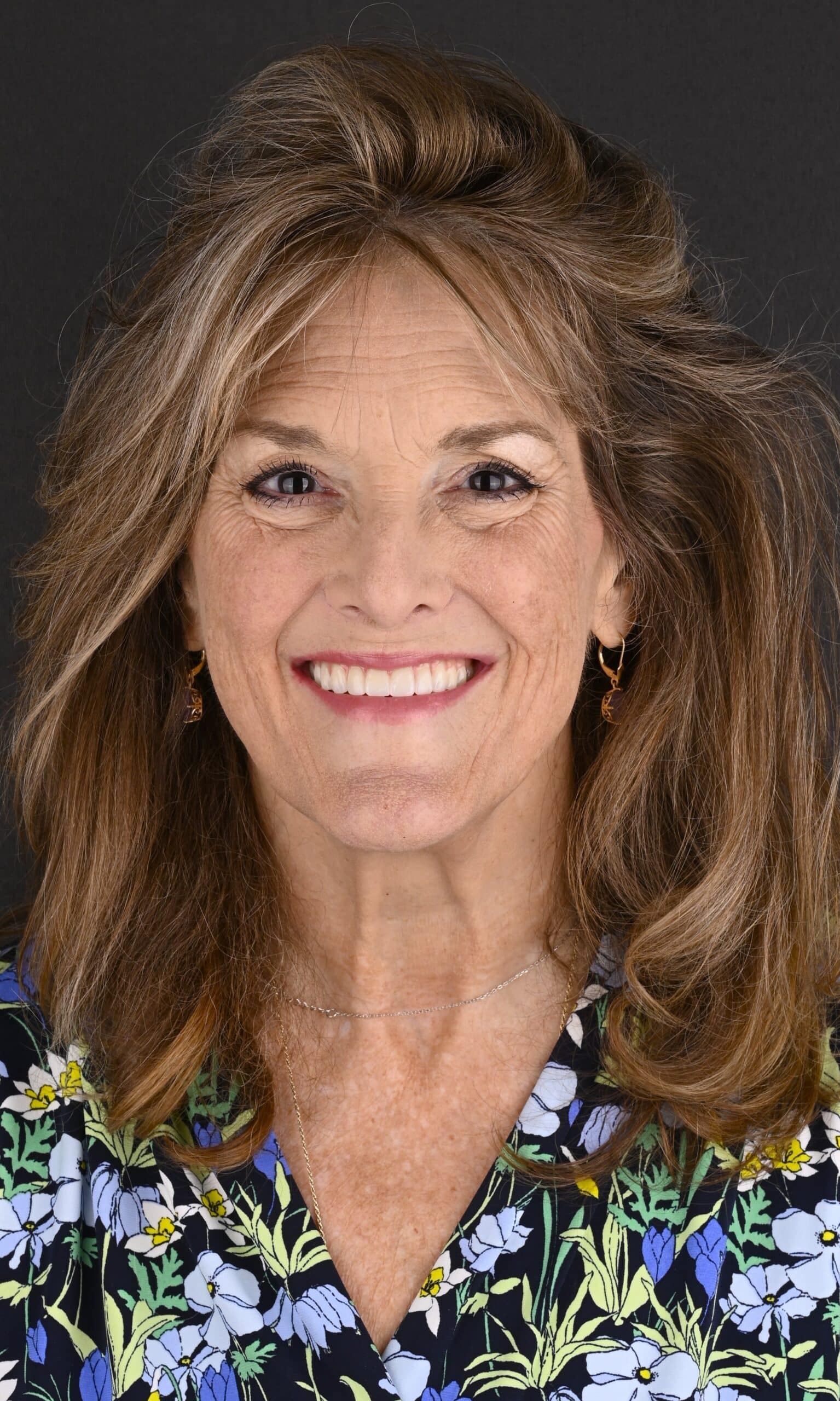 Smiling woman with long brown hair and a floral blouse against a dark background.