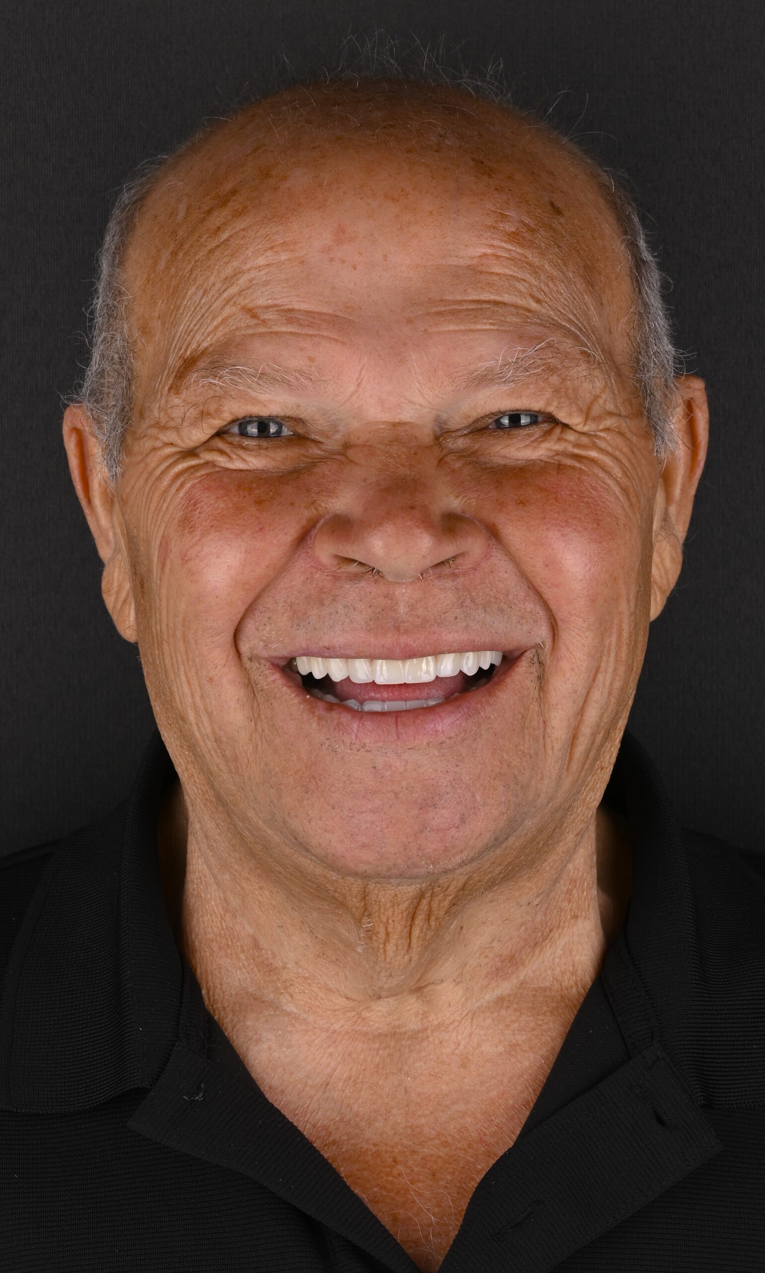 Close-up of an older man smiling broadly, with short gray hair and a black collared shirt, against a dark backdrop.