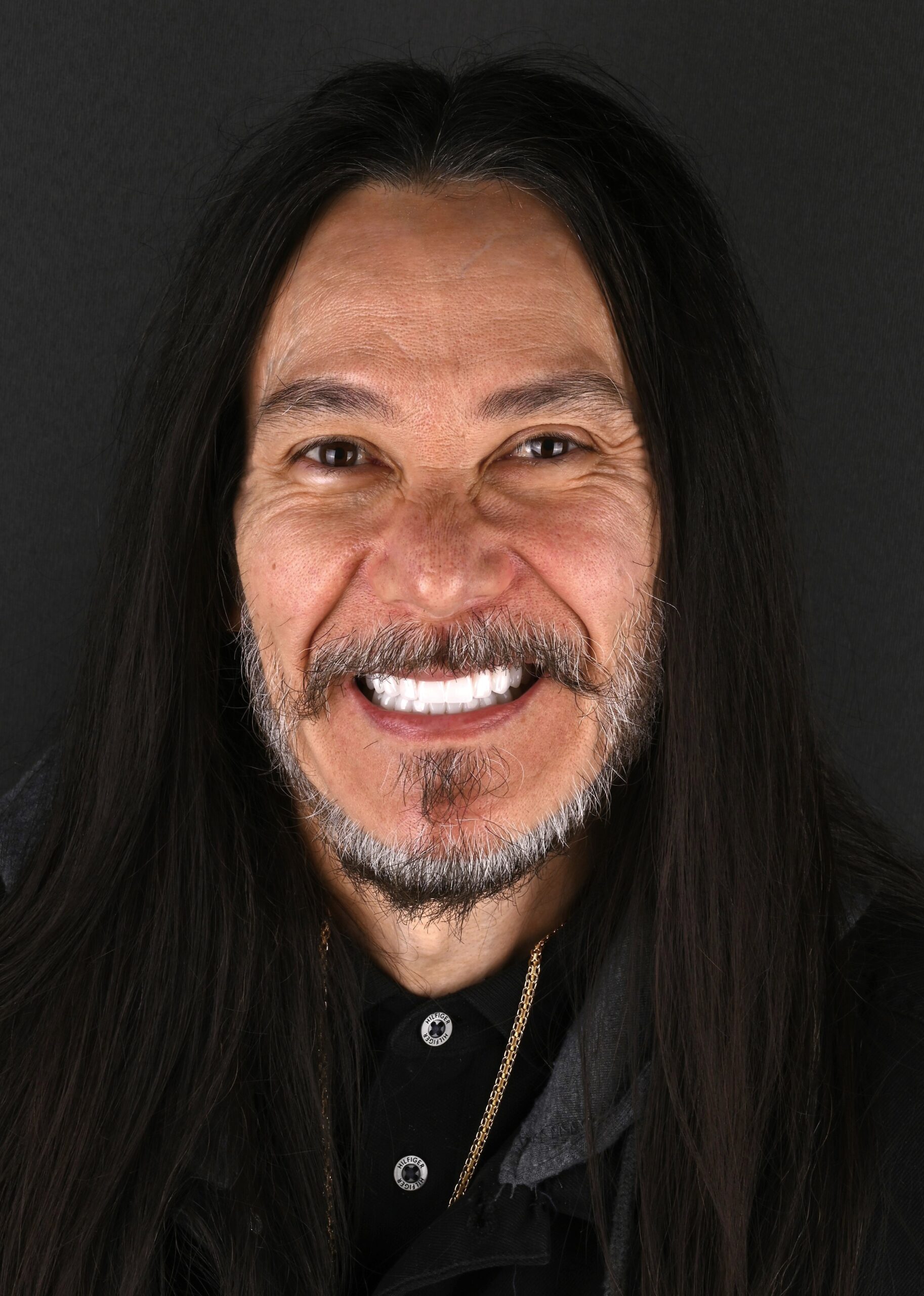 Smiling man with long dark hair and facial hair, wearing a black shirt against a dark background.