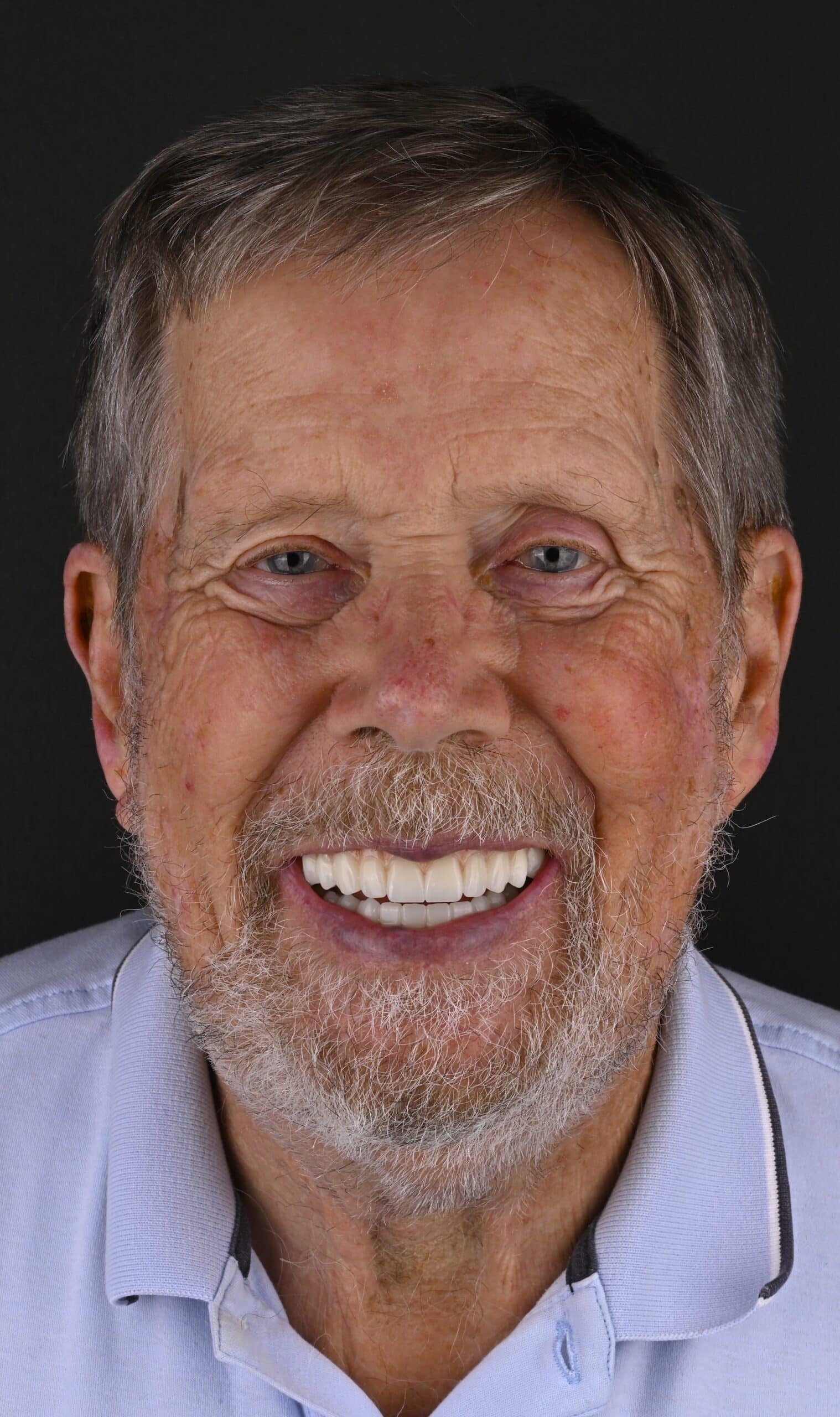 Close-up portrait of an older man with gray hair and beard, smiling widely against a dark background.