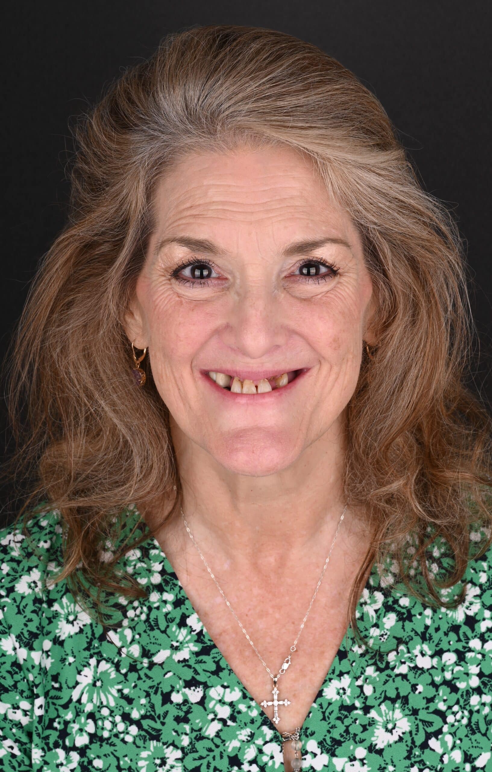 Smiling middle-aged woman with long brown hair wearing a green floral blouse and a silver cross necklace against a dark background.