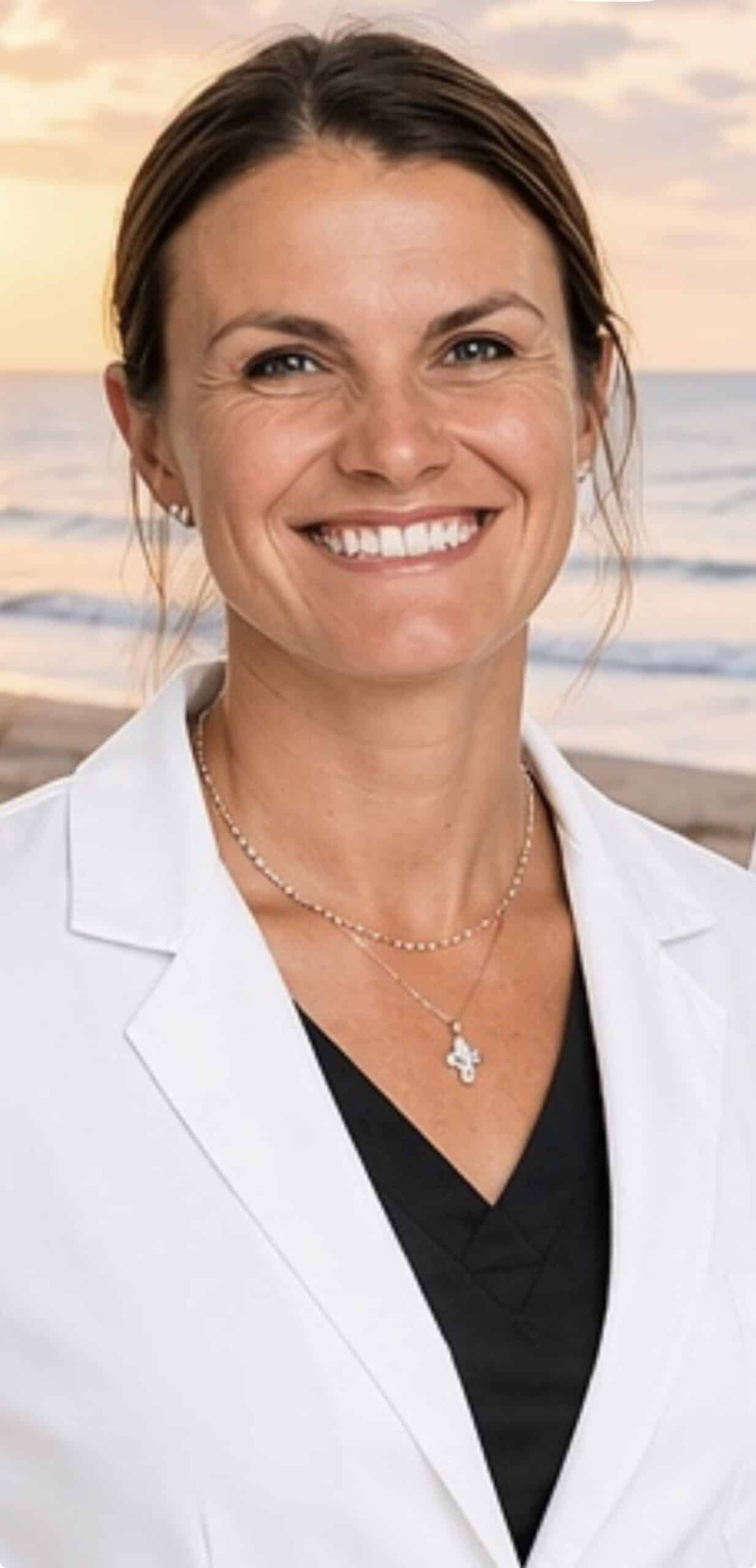 Smiling woman at the beach during sunset, wearing a white blazer and black top with delicate necklaces.