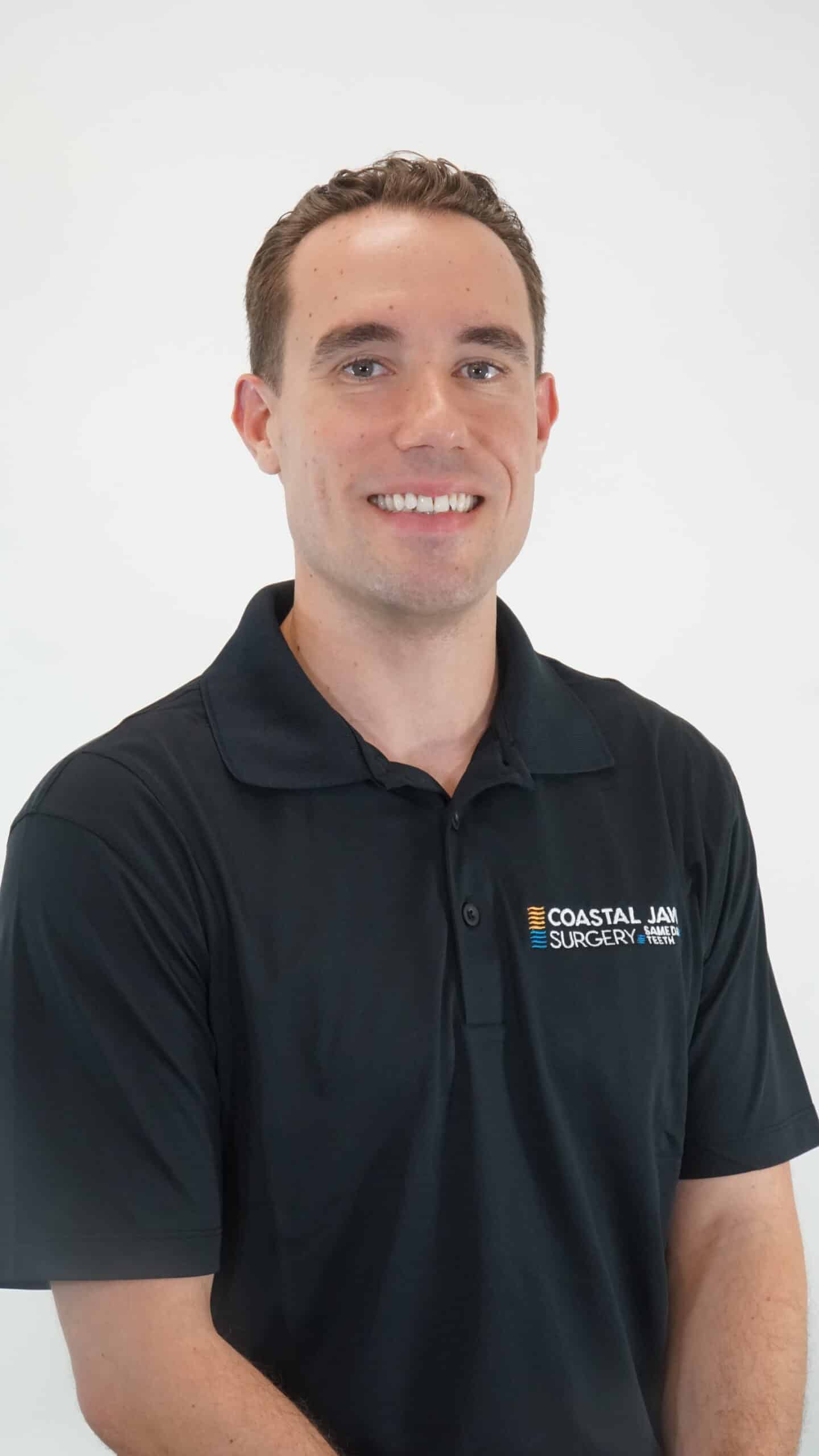 Smiling man in a black polo with Coastal Jaw Surgery logo against a white background.