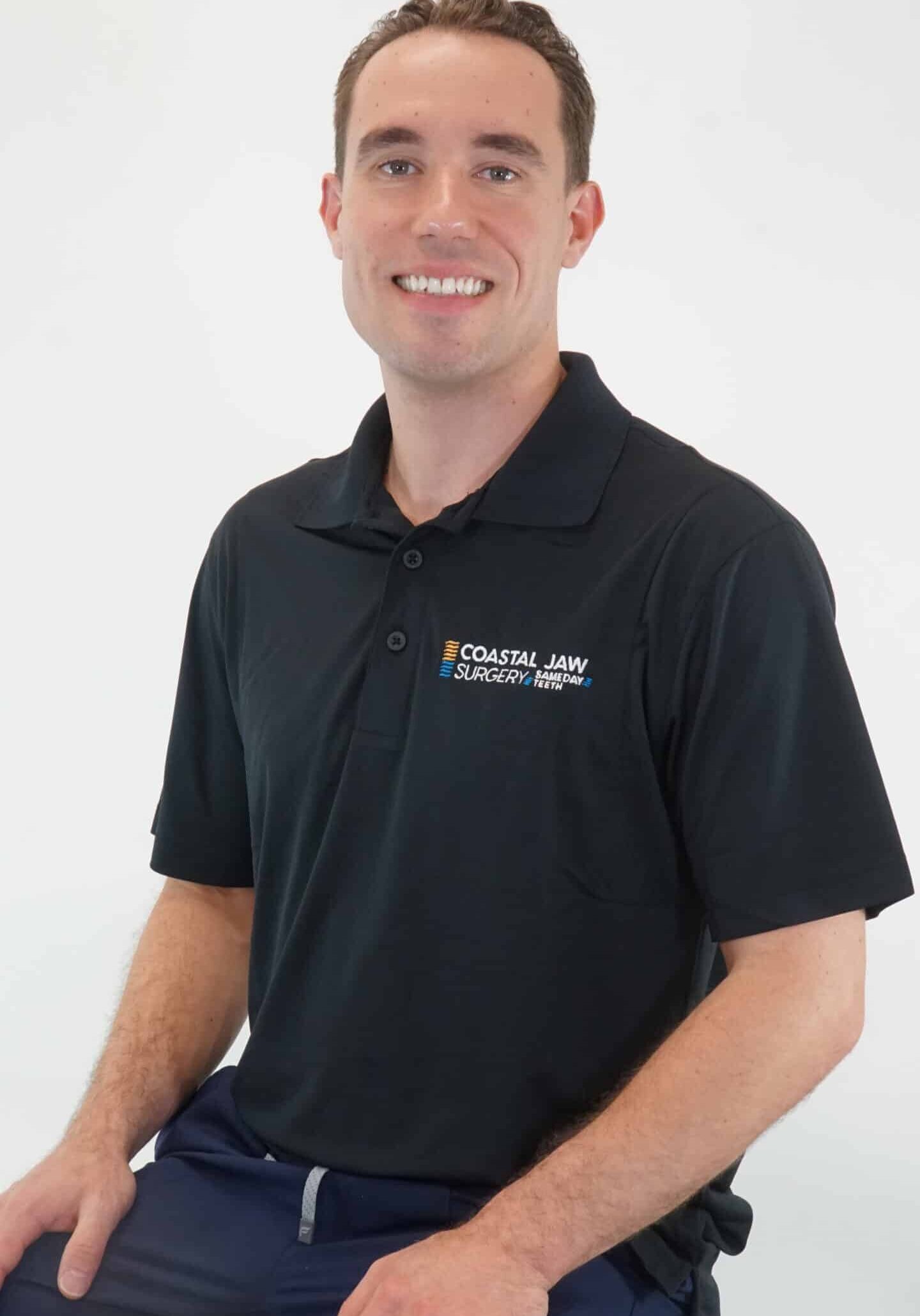 Smiling man in a black Coastal Jaw Surgery polo shirt, seated against a white background.